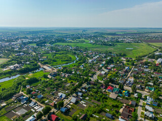 A wide aerial view of the city of Suzdal, showing a mix of residential houses, urban blocks, and a winding river with green fields in the distance.
