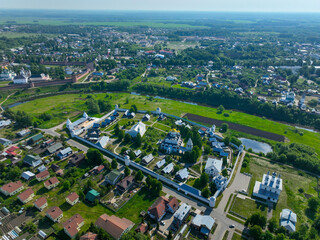 A wide aerial view of a monastery complex with white walls and golden domes, surrounded by residential houses and green fields in the city of Suzdal.