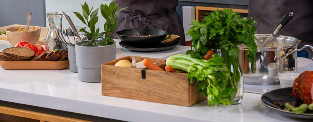 Fresh vegetables and herbs arranged on a kitchen countertop, with wooden box and utensils,...