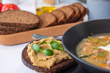 Freshly baked dark full grain bread with creamy hummus and vibrant greens served alongside a warm bowl of healthy soup, featuring nourishing ingredients and copy space