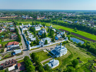A panoramic aerial view of the Pokrovsky Monastery complex and a separate, small white church, with residential houses and green fields in the background.