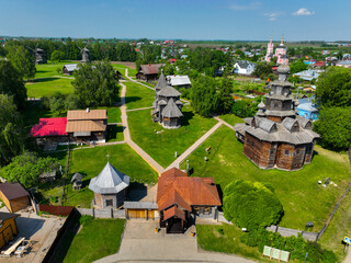 A high-angle aerial view of the Suzdal wooden architecture museum, showing various wooden buildings including windmills and a small house, with a town in the distance.