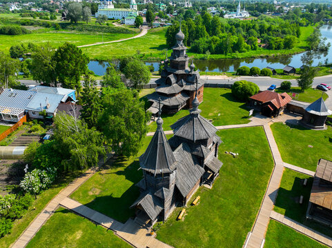 An aerial view of a historical wooden church with a bell tower, located next to a small river with residential houses and a modern town in the background.