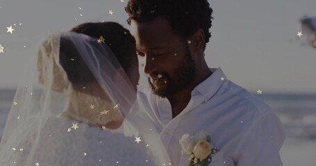 Facing bride and groom standing on sandy beach by ocean, with veil boutonniere and confetti overlay