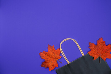 Black paper shopping bag and autumn leaves on an purple background. Autumn shopping and sales concept. Top view. Flatlay. Copy space.