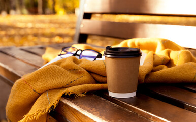 Takeaway cup of hot tasty coffee, warm blanket, eyeglasses and book on the bench in autumn park. Autumn cozy mood. Selective focus.