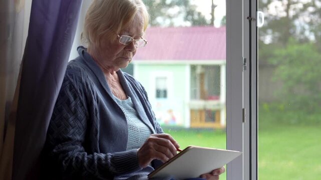 Elderly woman using digital tablet near window at home. Old female sitting on windowsill. Using apps or messengers. Living in nursing home or quarantine isolation