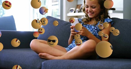 Girl in dress holding smartphone on grey couch at home, with floating 3D emoji spheres, bookshelf