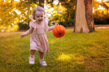 Cute two year old little girl with a bright smile standing in an autumn park, holding a small orange pumpkin in her hands. Warm sunlight, golden leaves in the background, atmospheric scene of autumn.