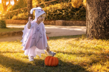 Smiling toddler in a white ghost costume with a pumpkin on the grass in an autumn park.
