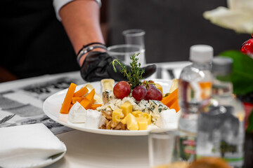 Assorted gourmet cheeses with grapes, walnuts, and thyme on a white plate, served by gloved hand in a formal dining setting. Close-up, vibrant food presentation.