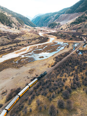 Aerial view of vintage train traveling through scenic mountain valley with winding river and autumn landscape