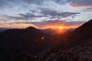 Scenic mountain sunset with glowing sun, dramatic sky, and soft clouds over rugged peaks in a peaceful natural landscape
