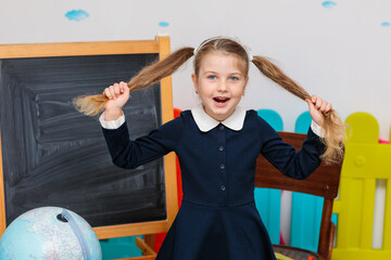 A playful girl in a blue school uniform pulls her ponytails and smiles brightly in front of a blackboard in a classroom.