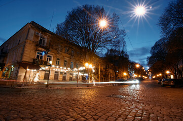 Fototapeta premium A charming nighttime view of a cobblestone street adorned with illuminated festive lighting, surrounded by historic architecture and tree-lined sidewalks, evoking a tranquil and nostalgic atmosphere.