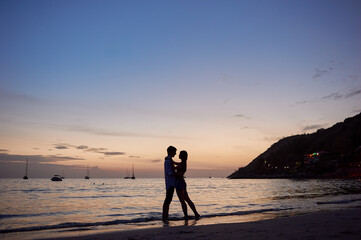 Naklejka premium Silhouetted couple shares a tender moment on a quiet beach during a breathtaking sunset, surrounded by calm waters and landscape, capturing love and tranquility in a scenic tropical destination.