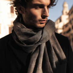 Mężczyzna z brązowymi włosami i zarostem, owinięty szalem w szaro-niebieskim kolorze. Patrzy w bok, na tle ulicy.