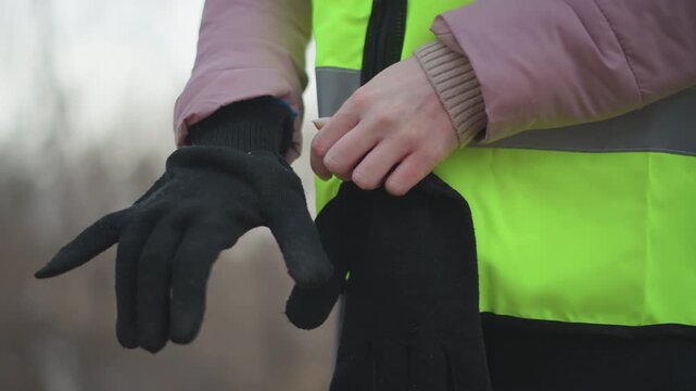 Close-up of person in neon yellow reflective safety vest and pink padded jacket putting on black knitted gloves outdoors during cold weather, preparing for outdoor work or safety task in winter