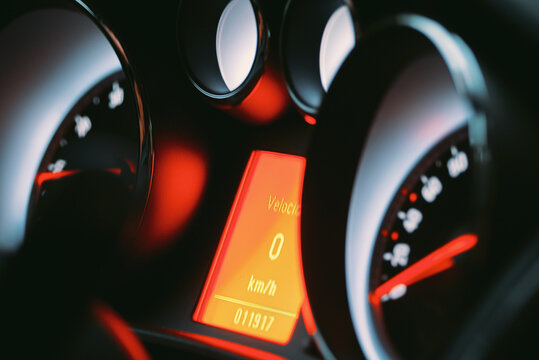 Close-up of a car's instrument cluster featuring a digital speed readout at zero kph with illuminated analog gauges and a red lighting theme