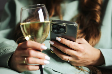 Woman holding wine glass and smartphone. Female hand with manicure multitasking. Relaxing indoors with technology. Drinking white wine while checking phone. Lifestyle with leisure and modern habits.