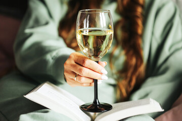Woman relaxing with wine glass closeup. Female hand with manicure holding wine. Reading book in soft green outfit. Evening cozy lifestyle indoors. Leisure, relaxation and quiet atmosphere.