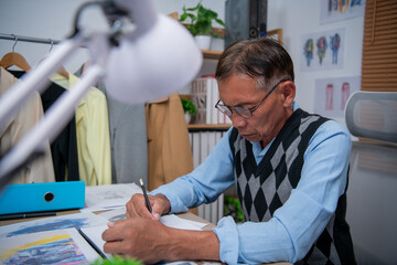 An elderly man in a sweater vest diligently sketches designs at his workspace.