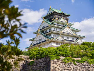 Osaka Castle viewed behind stone walls and green trees, with blue sky and clouds in Osaka, Japan.