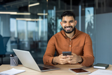 Smiling indian businessman sitting at an office desk, holding a smartphone while working with a laptop and tablet, portraying connectivity and professionalism