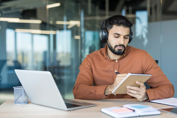 Young man with headphones works on a laptop in a modern glass-walled office, focused on taking notes in a notebook while studying or attending an online course for professional growth