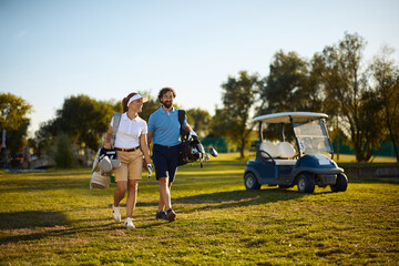 Fototapeta premium Happy couple of golfers carrying golf clubs in bags while walking on course.