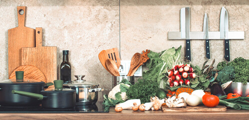 Kitchen countertop with cutting board, vegetables and utensils, pans on induction hob, trays etc. Cooking healthy food in the kitchen from local farm products