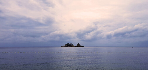 Tranquil seascape at sunset with a small rocky island covered in trees in the distance, surrounded by calm blue waters under a pastel sky.