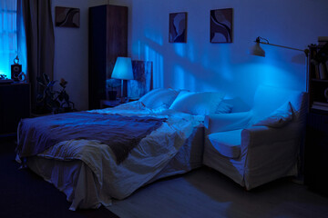 Empty bedroom with unmade bed and armchair illuminated by blue light, showing cozy nighttime setting with modern decor and soft shadows on walls and furniture © Mediaphotos