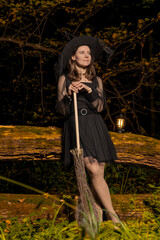 Woman in witch costume rests on broom next to lantern on a fallen forest log at night