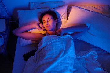 Woman lying in bed with eyes closed, smiling and resting head on pillow, holding blanket, smartphone visible beside, illuminated by soft blue and orange light © Mediaphotos