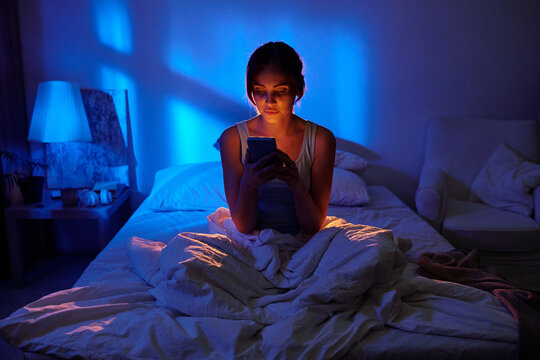 Woman sitting on bed using smartphone at night, illuminated by blue light from window, focused on screen, holding device with both hands, hair tied back