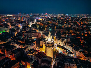 Aerial view of Galata Tower glowing above Beyoglu Istanbul Turkiye, crowded streets and warm lights spread through dense blocks, the skyline shimmers against deep blue night and vibrant urban energy