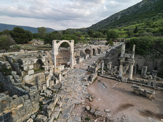 Stone arch frames the paved way in Ephesus, heavy blocks and fragments lie beside the walls, tourists explore the excavations, the legacy of ancient Turkey endures among marble and brick