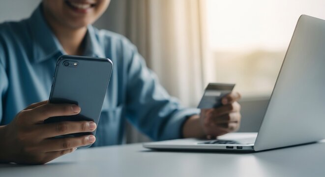 Smiling person uses smartphone and credit card for online shopping, laptop nearby - Powered by Adobe