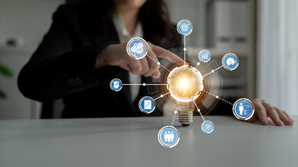 A professional businesswoman interacts with a glowing light bulb surrounded by digital icons. This...