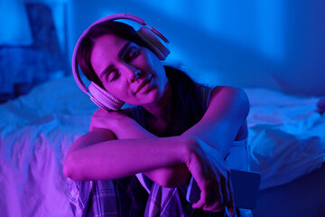 Woman sitting on floor with eyes closed, wearing headphones and holding smartphone, relaxing and listening to music in bedroom illuminated by blue and purple light