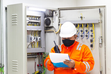 Electrician checking electrical panel safety while holding walkie-talkie and document, wearing PPE with hard hat and face mask ensuring protection against hazards and safety inspection protocols