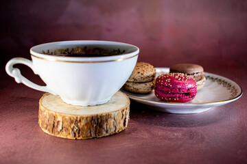 White porcelain cup with gold trim filled with dark coffee sits on a wooden coaster beside a plate of pink and brown macarons with sprinkles. Textured burgundy backdrop adds a cozy, elegant touch.