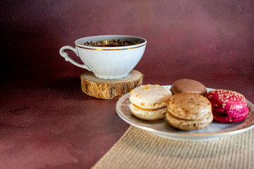 White porcelain cup with gold trim filled with dark coffee sits on a wooden coaster beside a plate of pink and brown macarons with sprinkles. Textured burgundy backdrop adds a cozy, elegant touch.