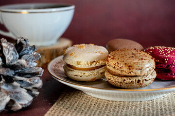 White cup of black coffee on a wooden coaster beside a pine cone and a plate of colorful macarons—white, brown, and pink—on a textured placemat. Dark background adds a warm, cozy ambiance.