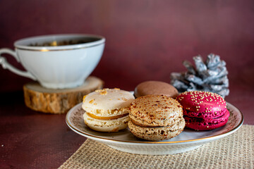 White cup of black coffee on a wooden coaster beside a pine cone and a plate of colorful macarons—white, brown, and pink—on a textured placemat. Dark background adds a warm, cozy ambiance.