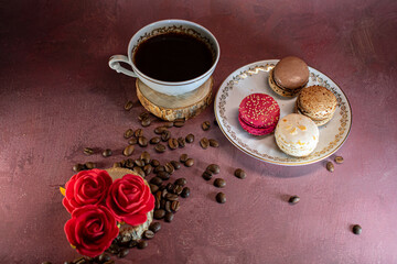 White coffee cup with gold trim on wooden coaster beside a plate of colorful macarons—chocolate, coffee, pink, and vanilla. Coffee beans scattered on a burgundy surface create a warm, inviting scene.