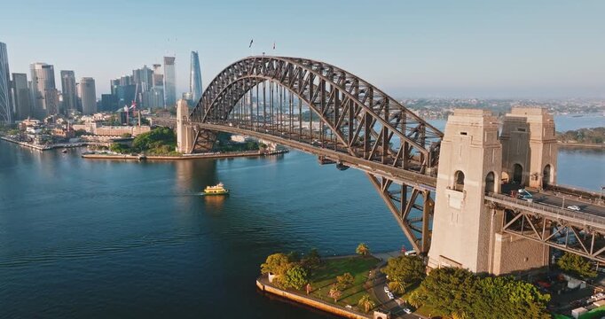 Australia, Sydney: Aerial view of Sydney Harbour Bridge with cars driving on it, connecting the city skyline with the surrounding landscape, on a sunny morning with blue sky. Drone flight footage - Powered by Adobe