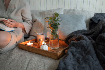 Cozy composition with woman in home wear reading book on the sofa with warm blanket, aromatic tea, candles and traditional autumn decorations close up. Hygge style composition.