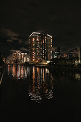 Fototapeta premium A luminous high-rise reflects its golden glow on a calm city river at night, surrounded by the dark, modern skyline.
